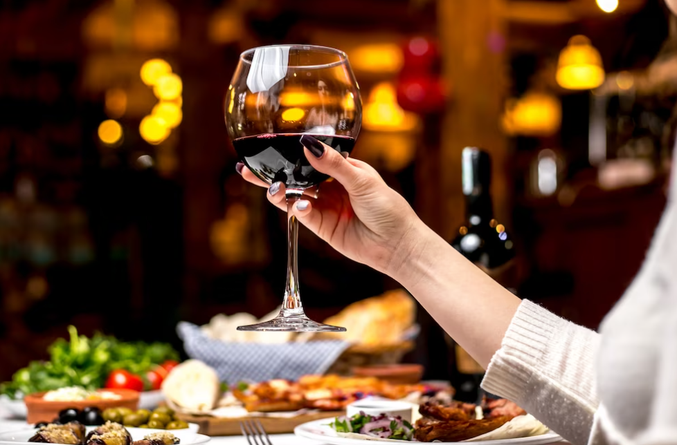 woman’s hand holding a wine glass with red wine in it, table with a bottle and plates with food