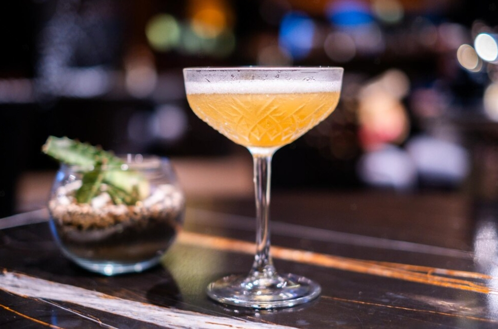 yellow cocktail in a  glass on the black table in the bar, flower in glass behind