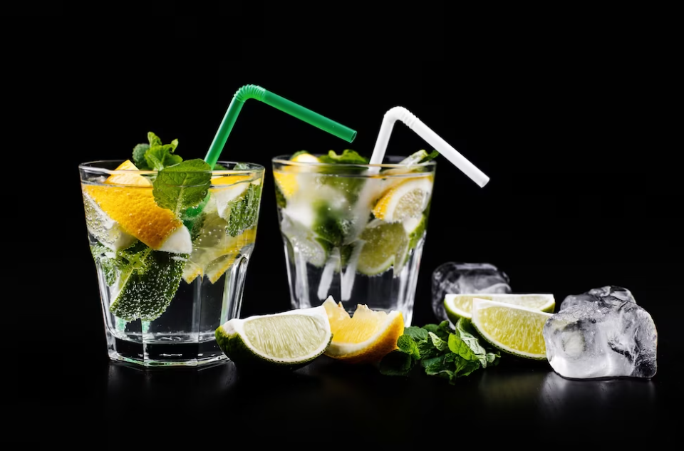 two glasses of cocktails with mint, lemon, straws, and ice on the black table